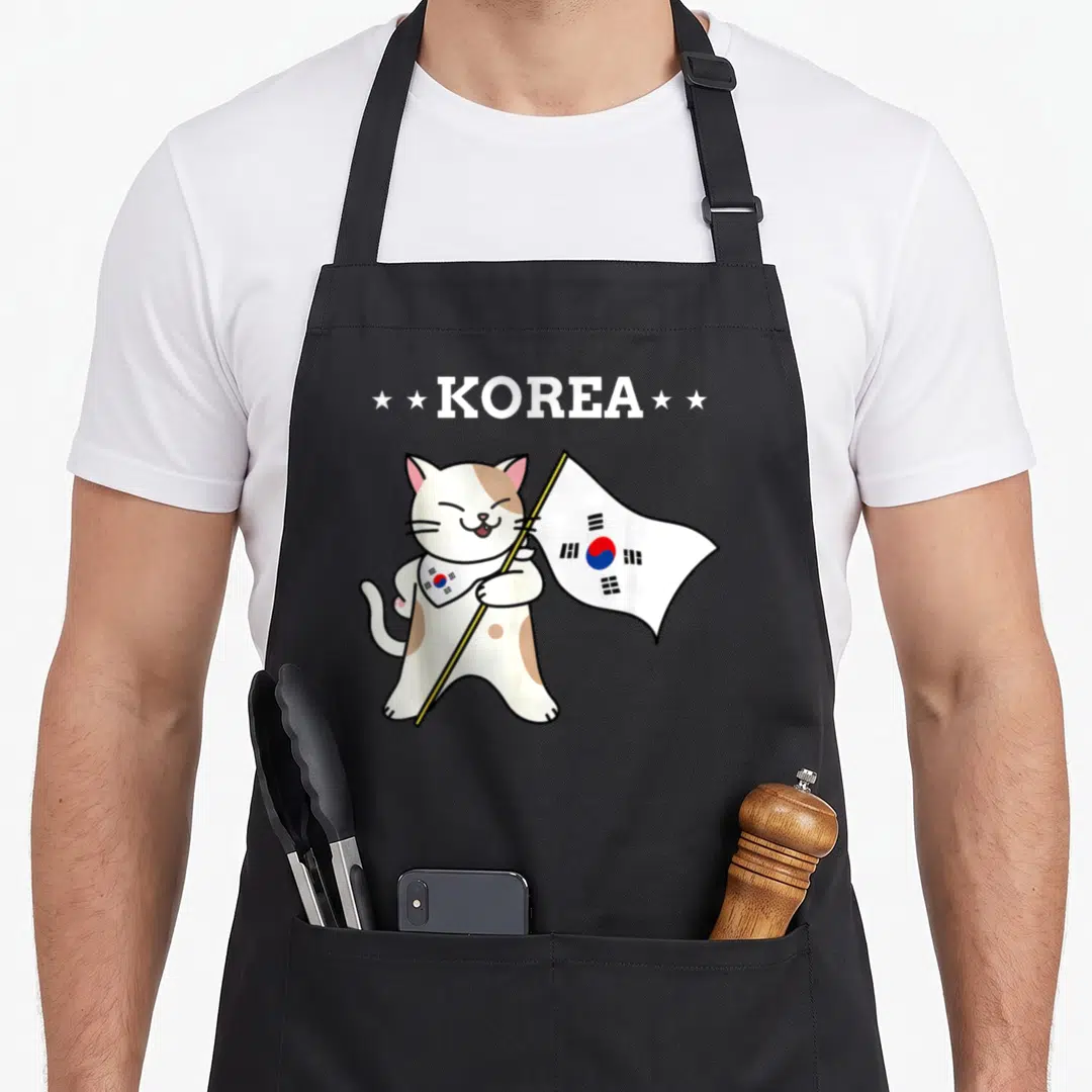 Korean cat apron - Cartoon cat holding KOREA flag with red blue and black details — shown on a black cotton canvas apron against a white background