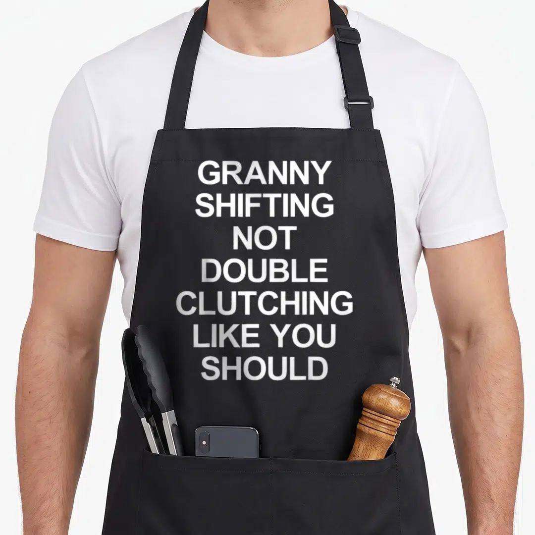 Grandma quick apron - White stacked text GRANNY SHIFTING NOT DOUBLE CLUTCHING LIKE YOU SHOULD on grey background — shown on a black cotton canvas apron against a white background
