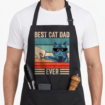Bump fist father day apron - Blue cat with pixelated sunglasses bumping a fist over retro stripes with BEST CAT DAD — shown on a black cotton canvas apron against a white background