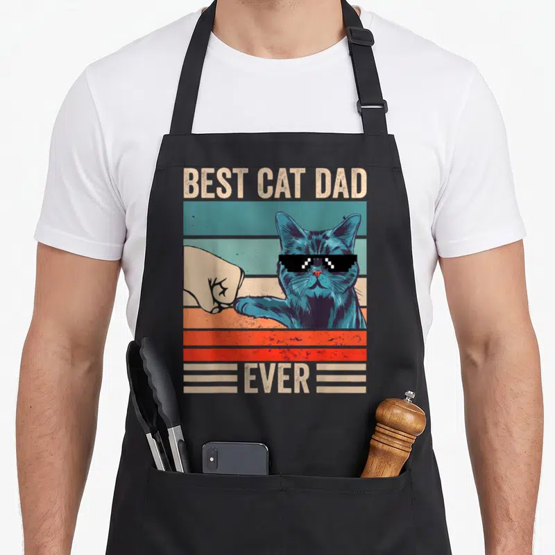 Bump fist father day apron - Blue cat with pixelated sunglasses bumping a fist over retro stripes with BEST CAT DAD — shown on a black cotton canvas apron against a white background
