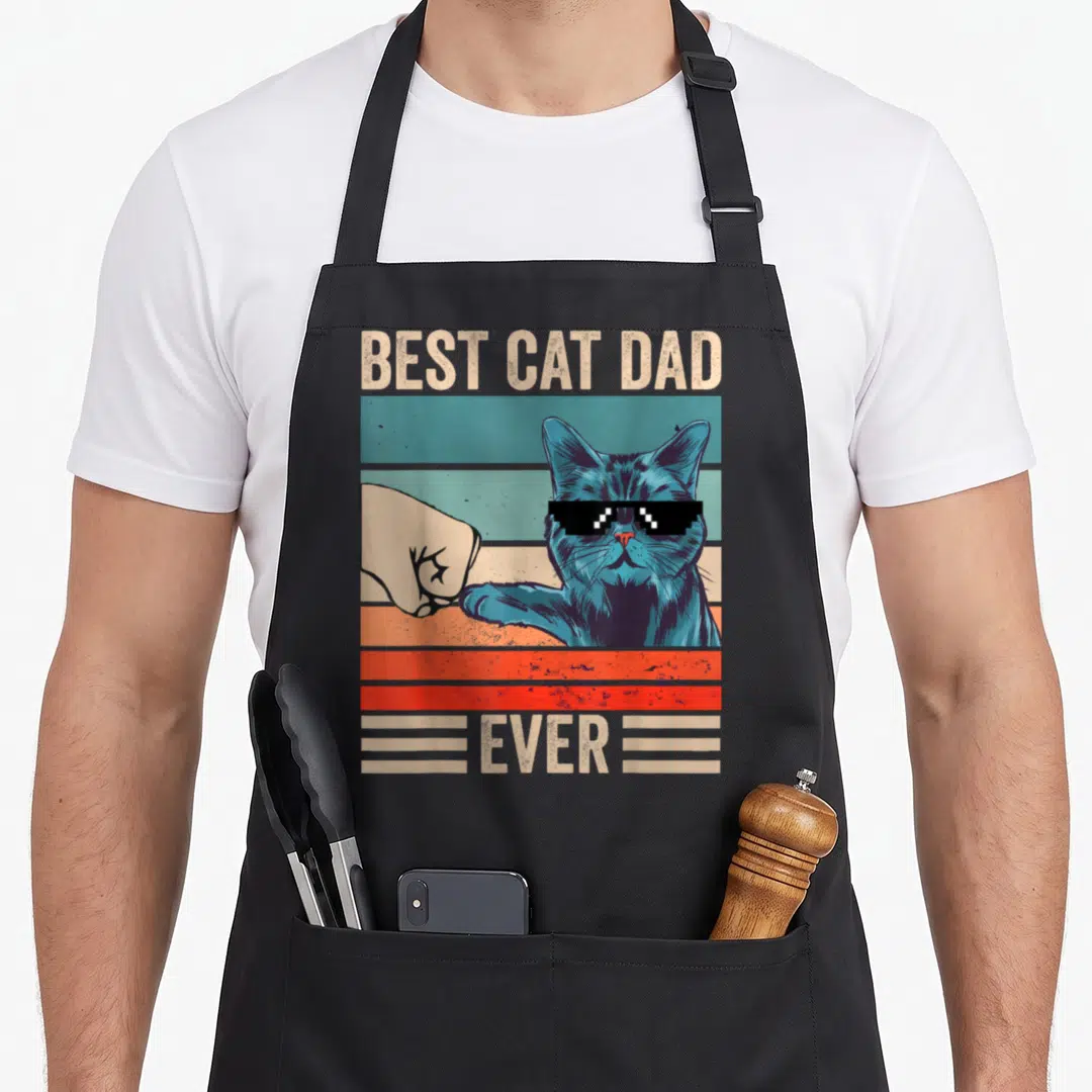 Bump fist father day apron - Blue cat with pixelated sunglasses bumping a fist over retro stripes with BEST CAT DAD — shown on a black cotton canvas apron against a white background