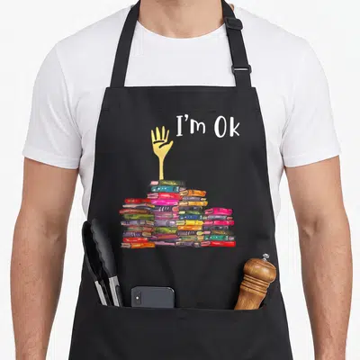 Boook funny book apron - Hand reaching from multicolored book pile with I'm Ok text in watercolor style — shown on a black cotton canvas apron against a white background