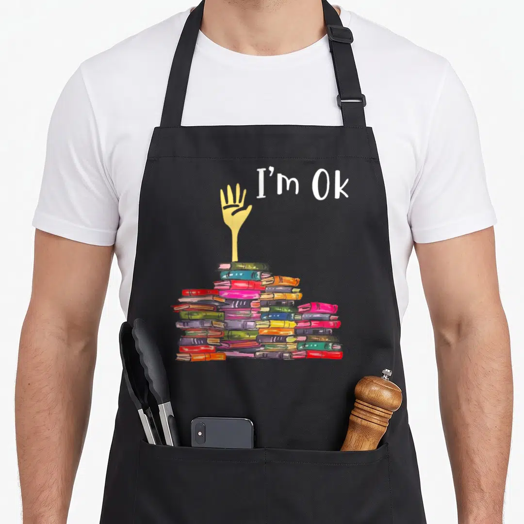 Boook funny book apron - Hand reaching from multicolored book pile with I'm Ok text in watercolor style — shown on a black cotton canvas apron against a white background