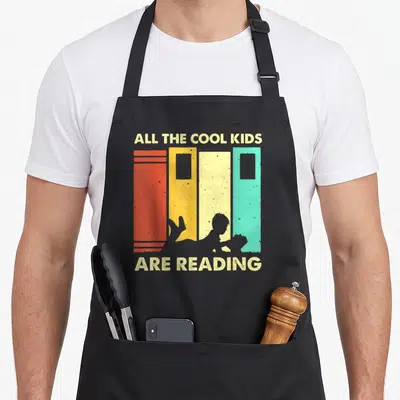 Reading book nerd apron - Retro graphic of child lying before four colorful books with text All The Cool Kids Are — shown on a black cotton canvas apron against a white background