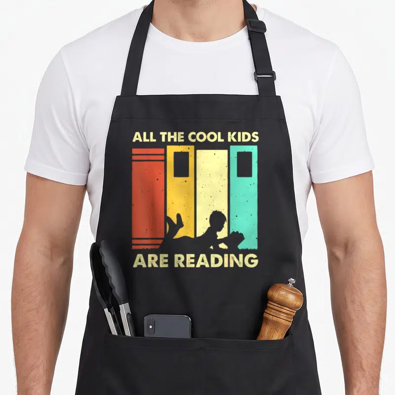 Reading book nerd apron - Retro graphic of child lying before four colorful books with text All The Cool Kids Are — shown on a black cotton canvas apron against a white background