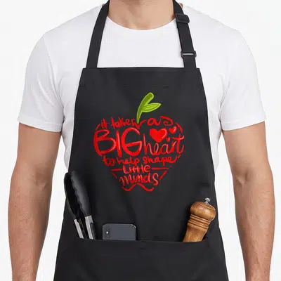 Appreciation apple teacher apron - Red text shaped like an apple with green leaf and scattered hearts containing — shown on a black cotton canvas apron against a white background