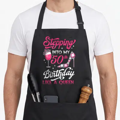 Wine birthday apron - Pink and white text Stepping Into My 50th Birthday Like A Queen with crowned wine glass — shown on a black cotton canvas apron against a white background