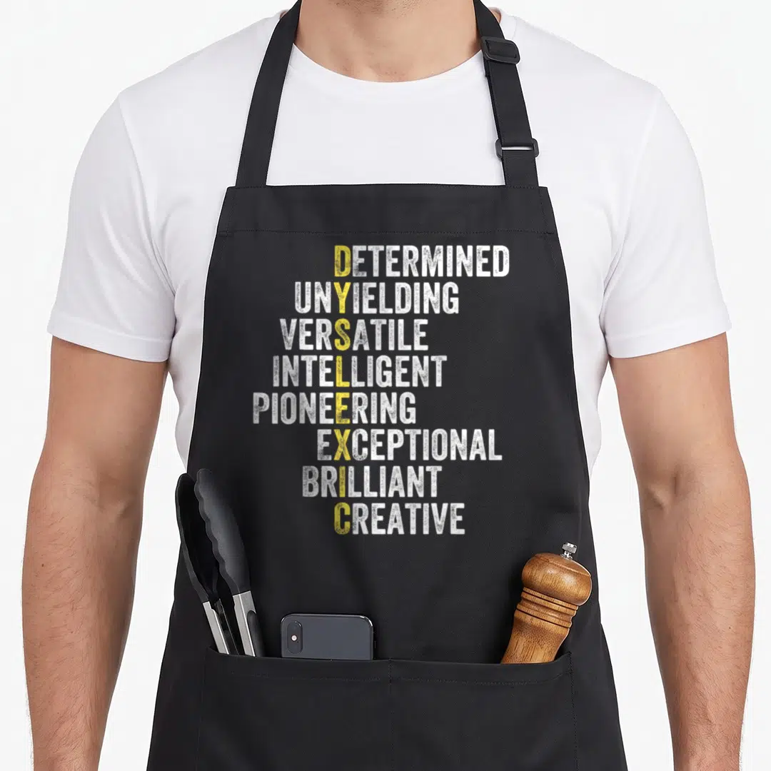Dyslexic my therapy apron - Stacked white and gold text reading DETERMINED UNYIELDING VERSATILE INTELLIGENT PIONEERING — shown on a black cotton canvas apron against a white background