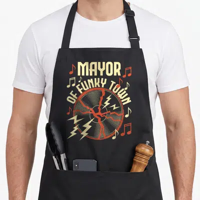 Groove funky apron - MAYOR OF FUNKY TOWN text over cracked vinyl record with red lightning bolts and musical notes — shown on a black cotton canvas apron against a white background