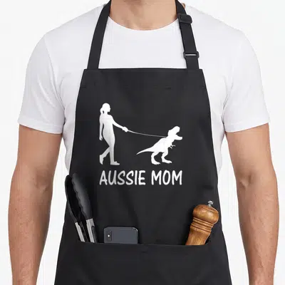 Dinosaur australian aprons - White silhouette of woman walking dinosaur on leash above AUSSIE MOM text — shown on a black cotton canvas apron against a white background