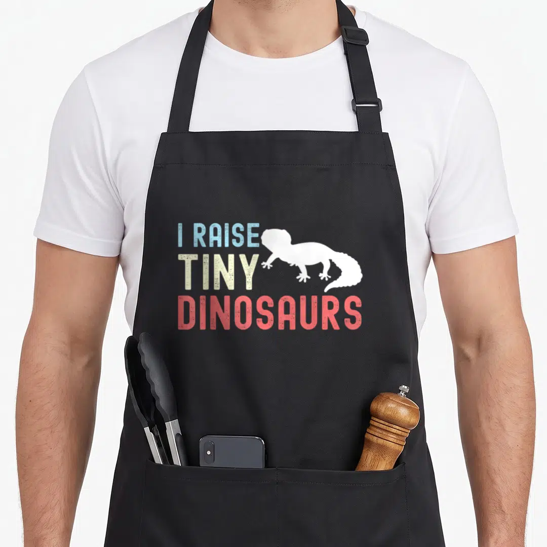 Raise leopard apron - Stacked I RAISE TINY DINOSAURS text beside white lizard silhouette in distressed blue yellow red — shown on a black cotton canvas apron against a white background