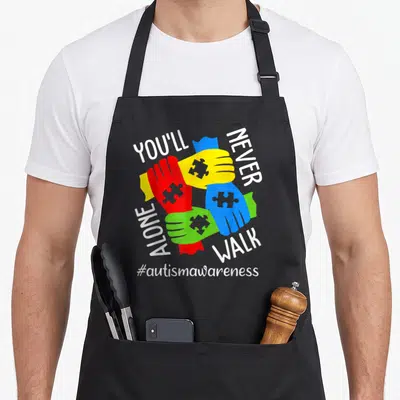 Autism youll never apron - Four interlocked hands in red yellow blue and green with puzzle piece details and awareness — shown on a black cotton canvas apron against a white background