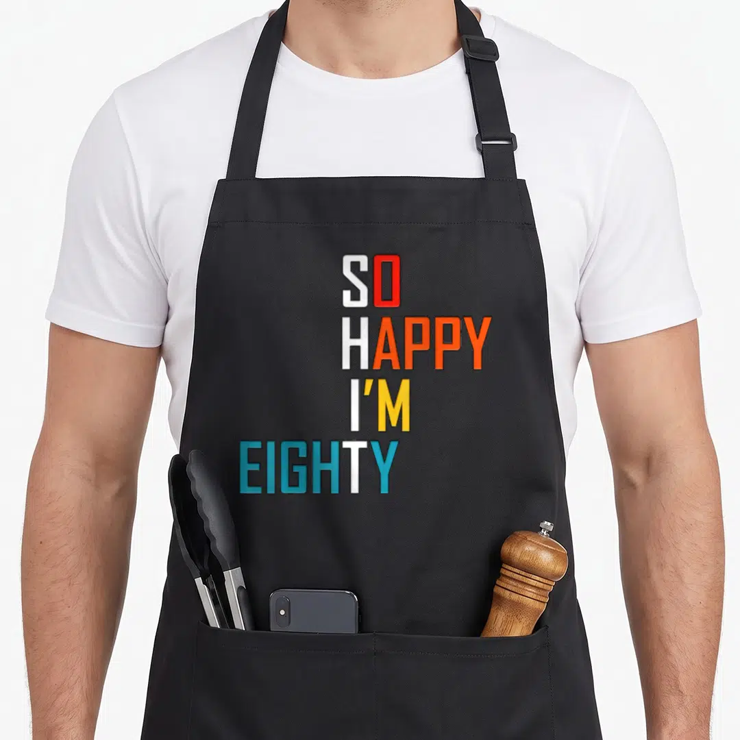 Eighty gag aprons - Stacked text SO HAPPY I'M EIGHTY in white red orange yellow blue — shown on a black cotton canvas apron against a white background
