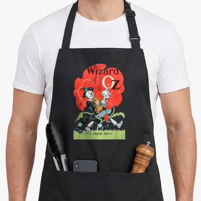Wizard dorothy apron - Vintage Wizard of Oz book cover with Scarecrow, Tin Man, and red poppy — shown on a black cotton canvas apron against a white background