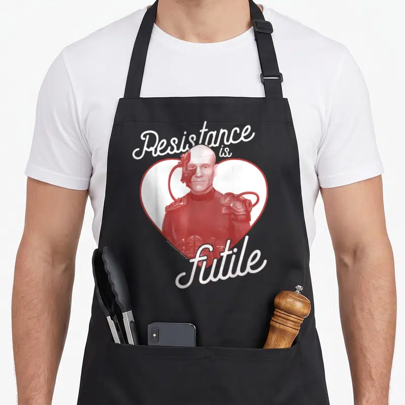 Generation valentine's day apron - Red Borg portrait in heart with white cursive text Resistance is futile — shown on a black cotton canvas apron against a white background