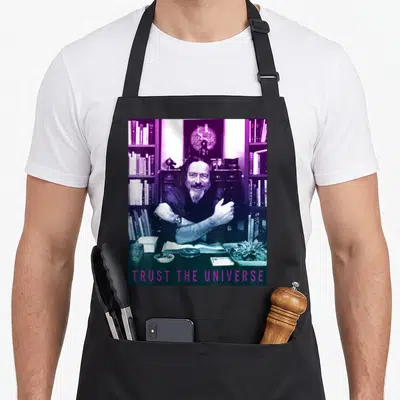 Philosopher famous aprons - Man at desk with bookshelves, TRUST THE UNIVERSE in purple text — shown on a black cotton canvas apron against a white background