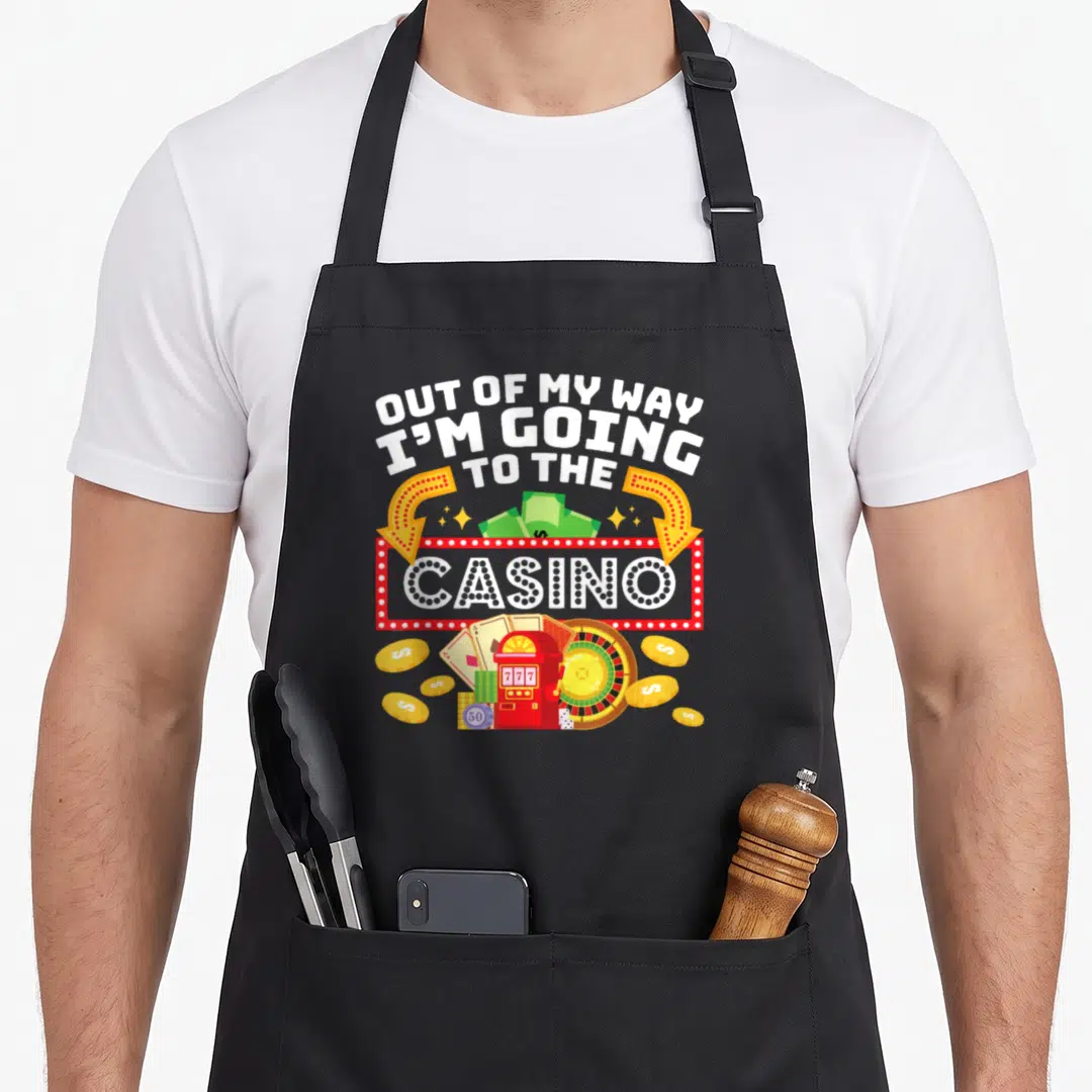 Way casino aprons - OUT OF MY WAY text above red marquee sign, slot machine, roulette wheel, and gold coins — shown on a black cotton canvas apron against a white background