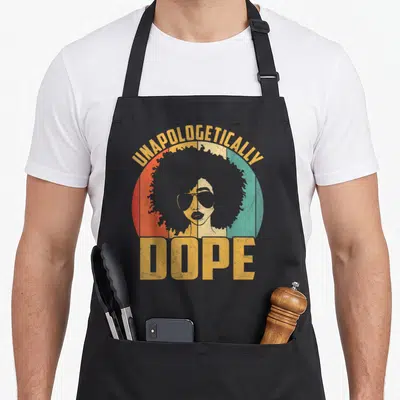 Unapologetically african print apron - Retro woman with afro and sunglasses in red green yellow circle with text — shown on a black cotton canvas apron against a white background