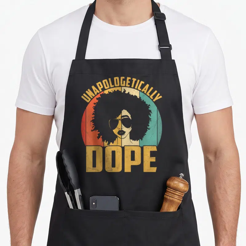 Unapologetically african print apron - Retro woman with afro and sunglasses in red green yellow circle with text — shown on a black cotton canvas apron against a white background