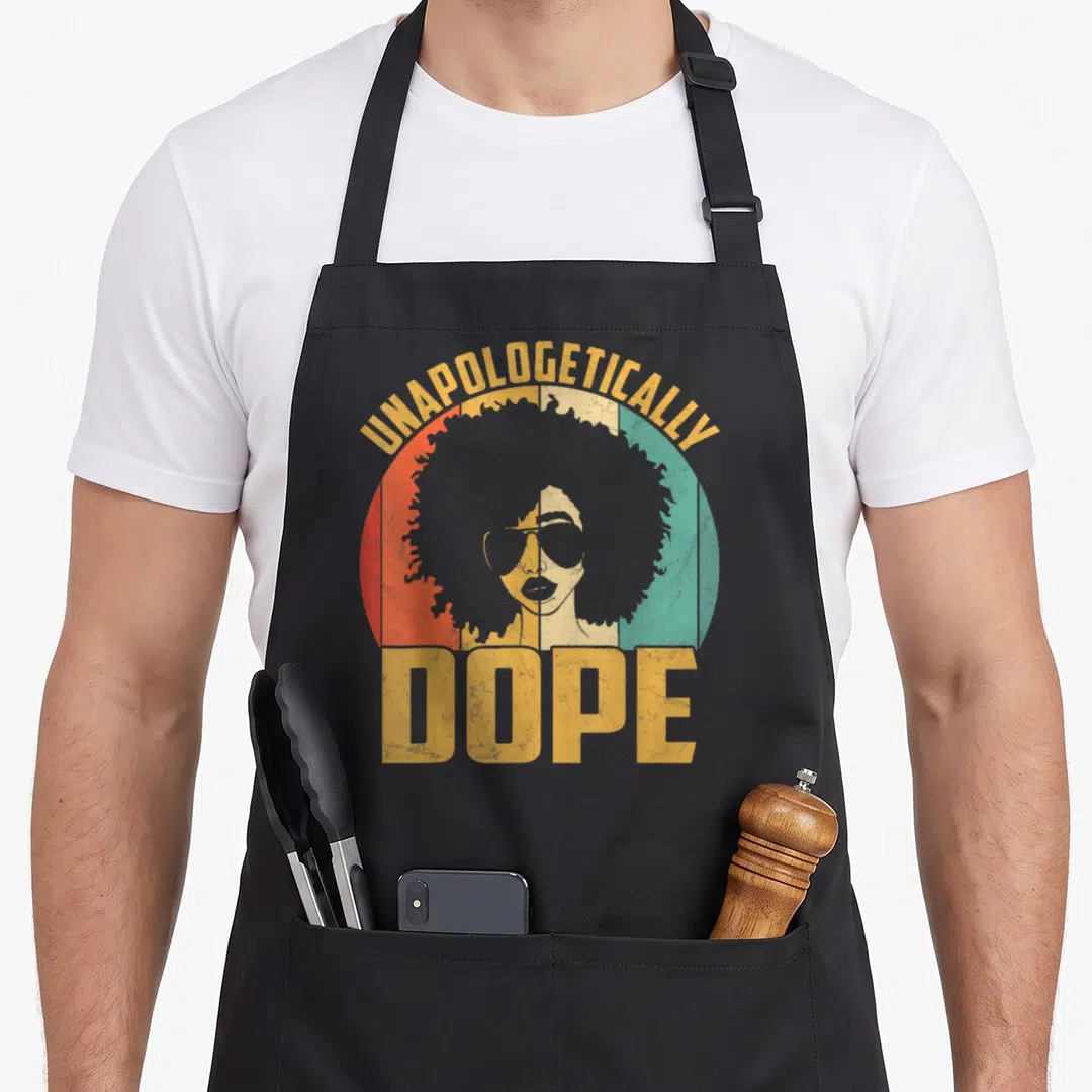Unapologetically african print apron - Retro woman with afro and sunglasses in red green yellow circle with text — shown on a black cotton canvas apron against a white background
