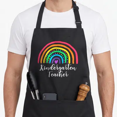 Funny rainbow kindergarten teacher apron - Cartoon rainbow with purple heart and stacked white Kindergarten Teacher text — shown on a black cotton canvas apron against a white background