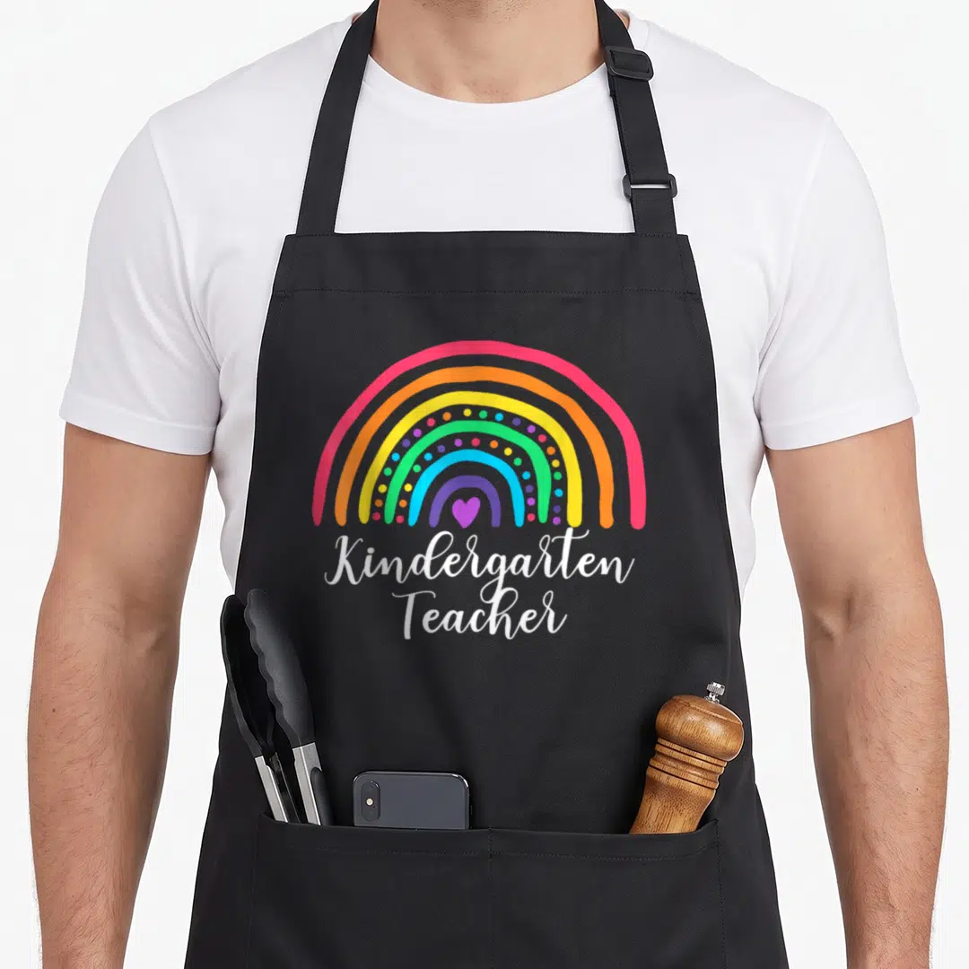 Funny rainbow kindergarten teacher apron - Cartoon rainbow with purple heart and stacked white Kindergarten Teacher text — shown on a black cotton canvas apron against a white background