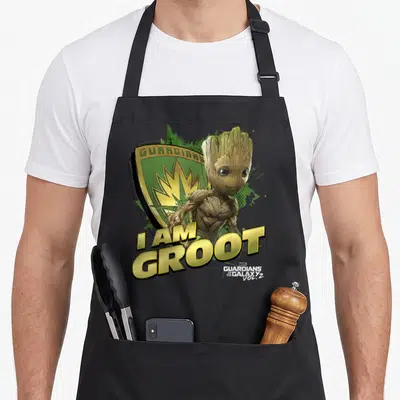 Galaxy defender apron - I AM GROOT text below photo-style Groot figure and green gold shield logo — shown on a black cotton canvas apron against a white background