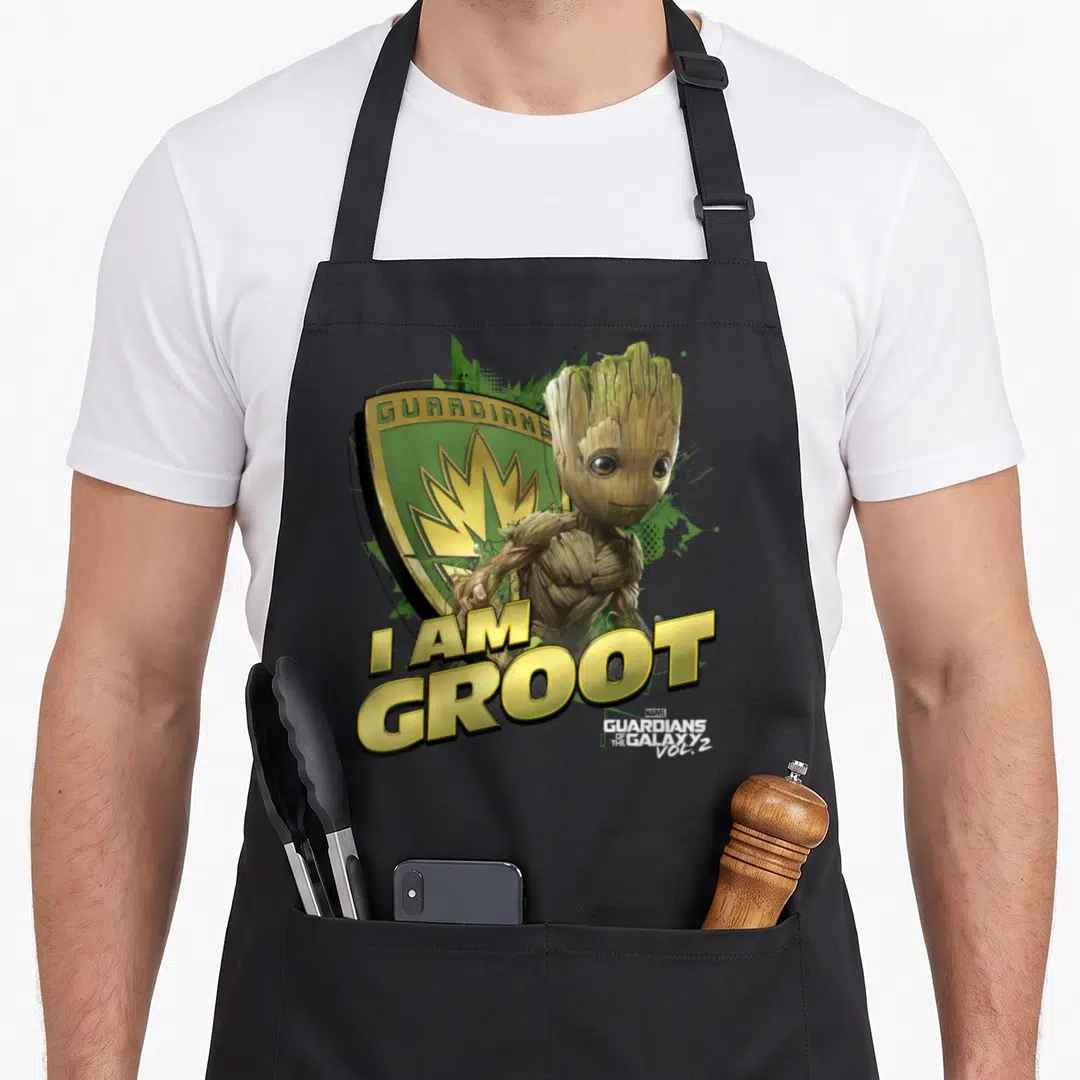 Galaxy defender apron - I AM GROOT text below photo-style Groot figure and green gold shield logo — shown on a black cotton canvas apron against a white background