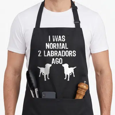 Normal labrador apron - White text I WAS NORMAL 2 LABRADORS AGO above two facing dog silhouettes — shown on a black cotton canvas apron against a white background