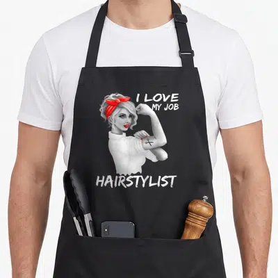 Salon hairstylist apron - Woman flexing arm with scissors tattoo and I LOVE MY JOB HAIRSTYLIST text — shown on a black cotton canvas apron against a white background