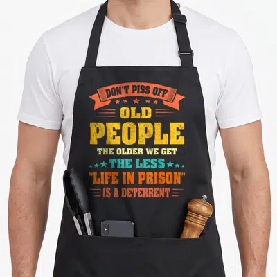 Older novelty apron - Distressed text reading Don't Piss Off Old People Life In Prison deterrent in yellow orange — shown on a black cotton canvas apron against a white background