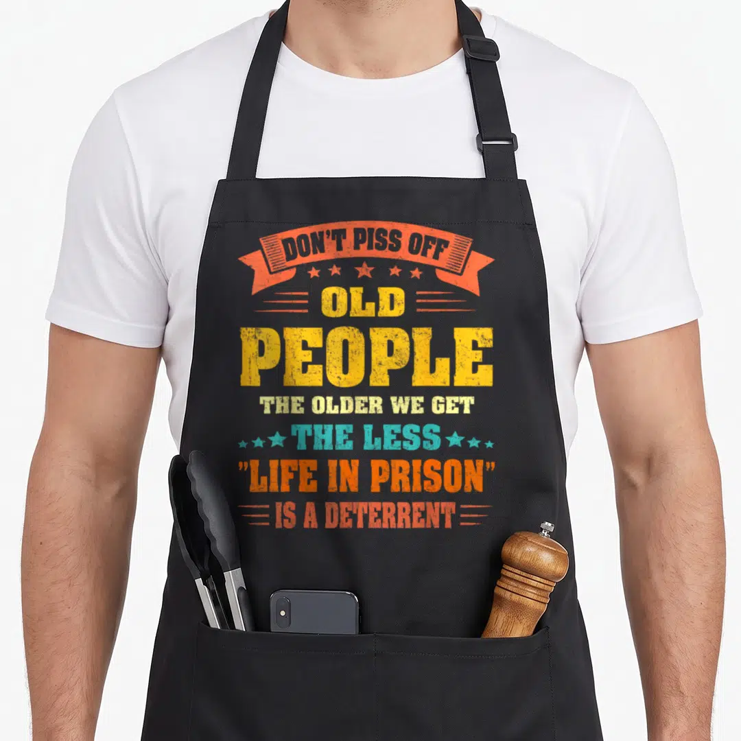 Older novelty apron - Distressed text reading Don't Piss Off Old People Life In Prison deterrent in yellow orange — shown on a black cotton canvas apron against a white background