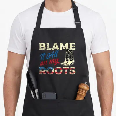 Blame southern aprons - Stacked BLAME It All on my ROOTS text with cowboy boots and flag pattern fill — shown on a black cotton canvas apron against a white background