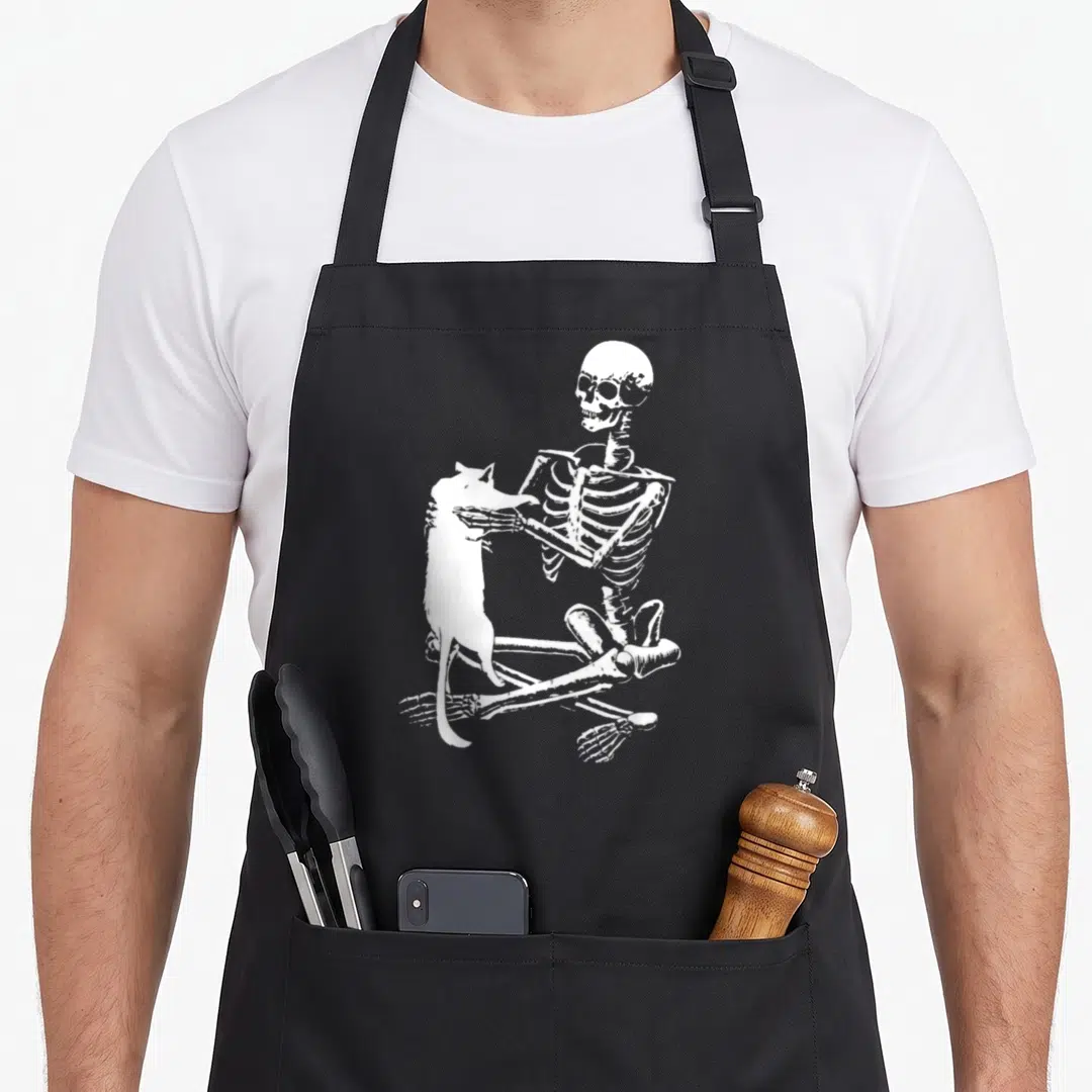 Holding funny halloween aprons - White line art of a skeleton sitting cross-legged petting a standing cat — shown on a black cotton canvas apron against a white background