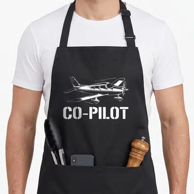 Aircraft pilot chef and co aprons - White line-art airplane above CO-PILOT stencil text on dark fabric — shown on a black cotton canvas apron against a white background