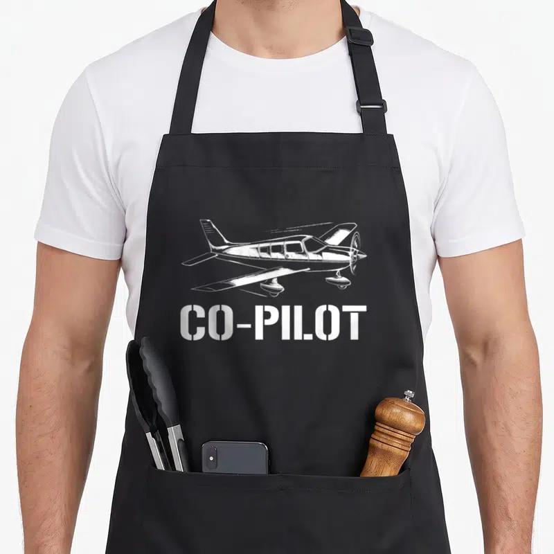 Aircraft pilot chef and co aprons - White line-art airplane above CO-PILOT stencil text on dark fabric — shown on a black cotton canvas apron against a white background