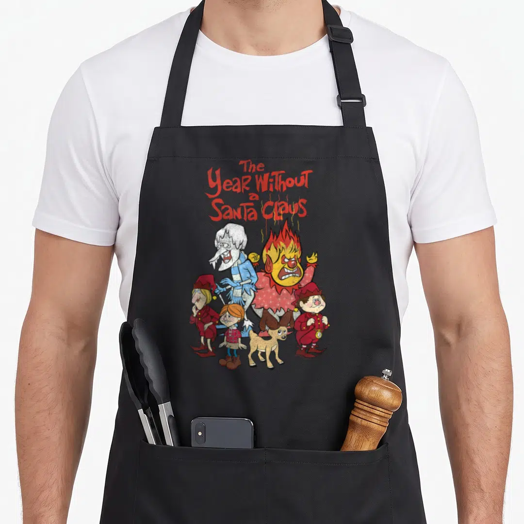 Shot santa claus apron - Red stacked text The Year Without a Santa Claus above Heat Miser Snow Miser and elves — shown on a black cotton canvas apron against a white background