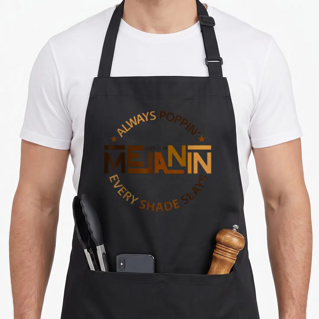 Beauty magic apron - Brown and orange circular text reading MADE WITH MELANIN with two stars flanking the center — shown on a black cotton canvas apron against a white background