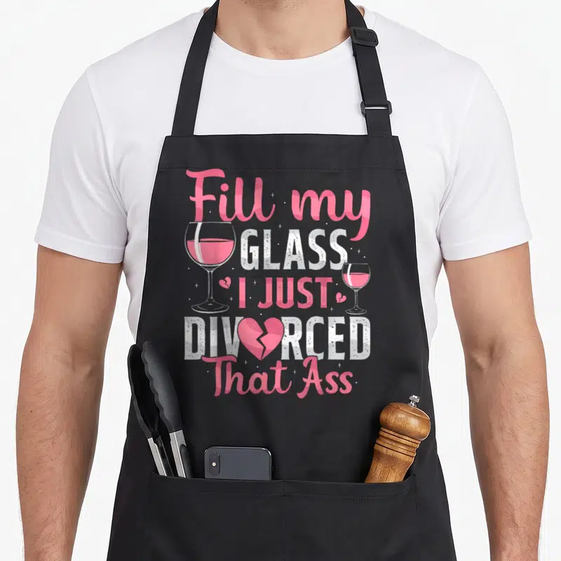 Fill divorce party apron - Pink and white stacked text reading Fill My Glass I Just Divorced That Ass flanked by two — shown on a black cotton canvas apron against a white background