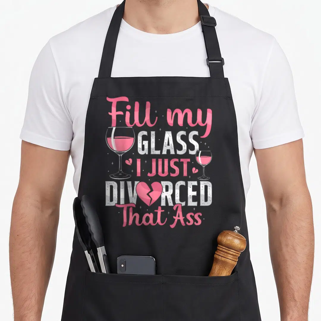 Fill divorce party apron - Pink and white stacked text reading Fill My Glass I Just Divorced That Ass flanked by two — shown on a black cotton canvas apron against a white background