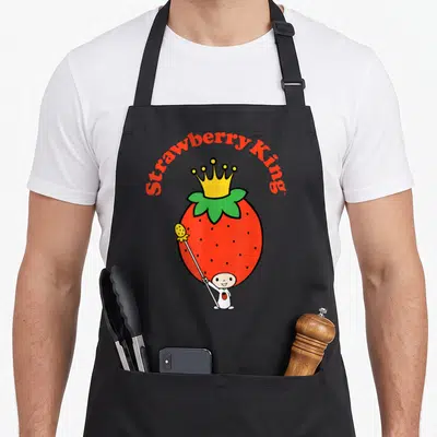 King strawberry apron - Red strawberry with yellow crown and small figure holding scepter under arched text — shown on a black cotton canvas apron against a white background