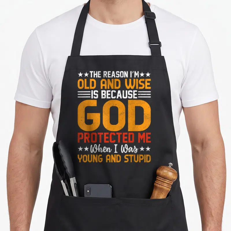 Reason wise apron - Stacked distressed text reading THE REASON I'M OLD AND WISE in yellow white and red with stars — shown on a black cotton canvas apron against a white background