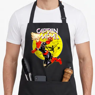 Earth captain apron - Captain Marvel leaping in front of yellow circle and black plane with bold text above — shown on a black cotton canvas apron against a white background