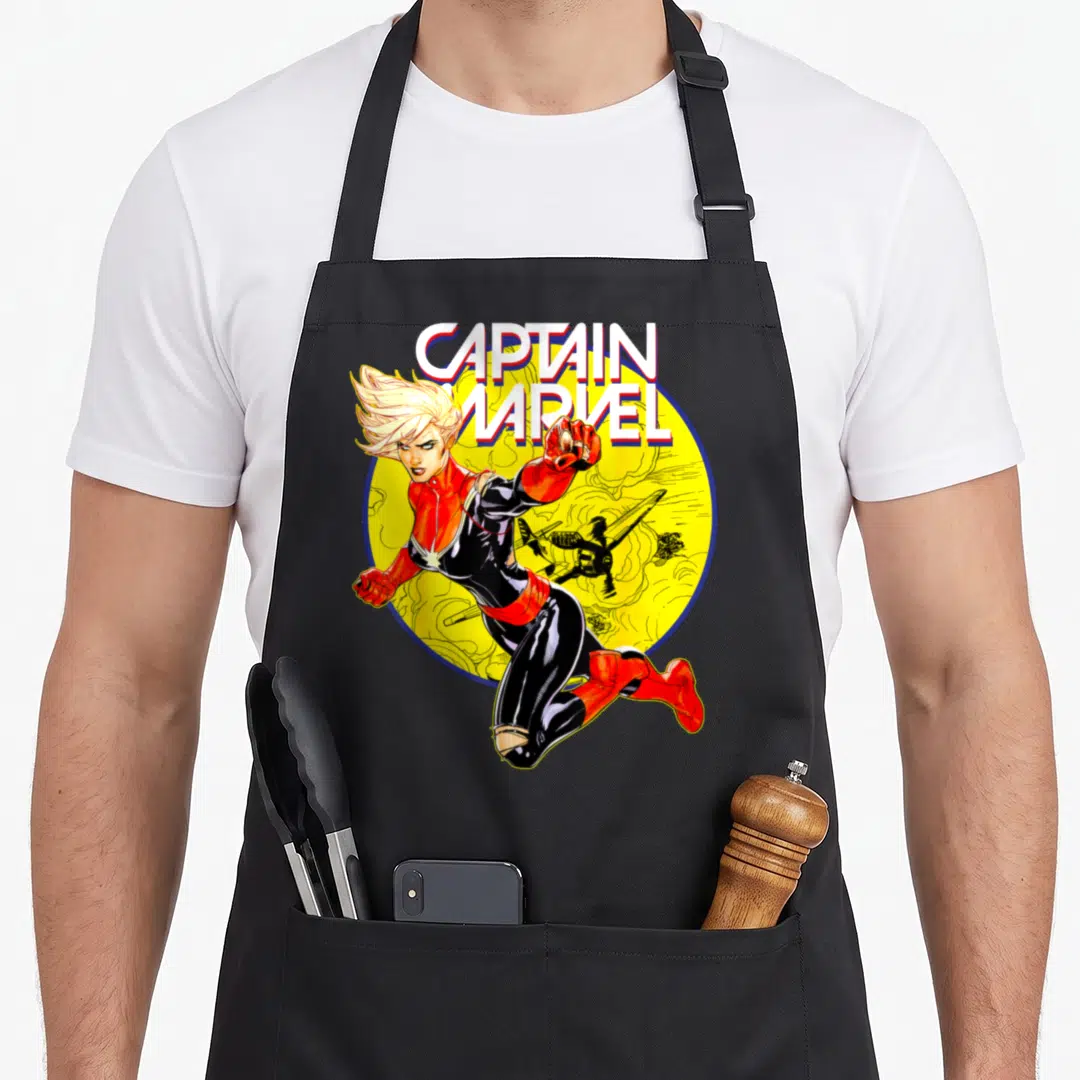 Earth captain apron - Captain Marvel leaping in front of yellow circle and black plane with bold text above — shown on a black cotton canvas apron against a white background