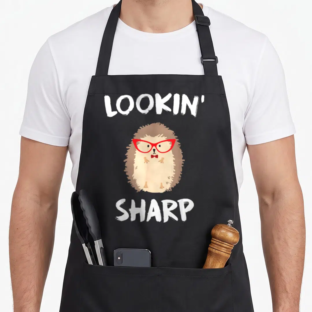 Cool hedgehog mom apron - Cartoon hedgehog with red glasses and bow tie between white text LOOKIN SHARP — shown on a black cotton canvas apron against a white background