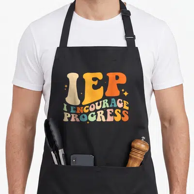 Education school teacher apron - Retro text print reading IEP I Encourage Progress in orange beige blue and pink — shown on a black cotton canvas apron against a white background