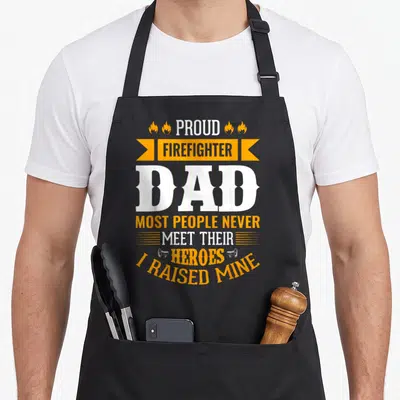 Meet firefighter apron - Stacked white and orange text reading PROUD FIREFIGHTER DAD with flames and megaphone icons — shown on a black cotton canvas apron against a white background