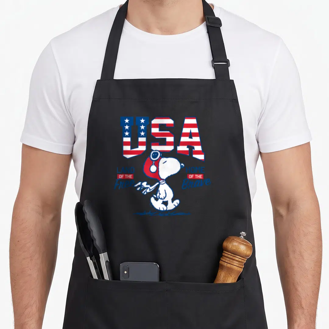 Free july usa apron - Snoopy in aviator hat under USA flag letters with Land of the Free Home of the Brave text — shown on a black cotton canvas apron against a white background