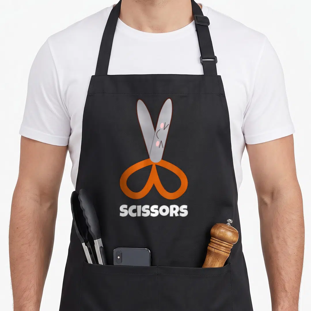 Rock scissor paper apron - Cartoon pair of scissors with orange handles and face above the word SCISSORS — shown on a black cotton canvas apron against a white background