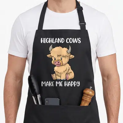 Messy highland cow apron - Cartoon tan sitting highland cow with white text reading HIGHLAND COWS MAKE ME HAPPY above — shown on a black cotton canvas apron against a white background